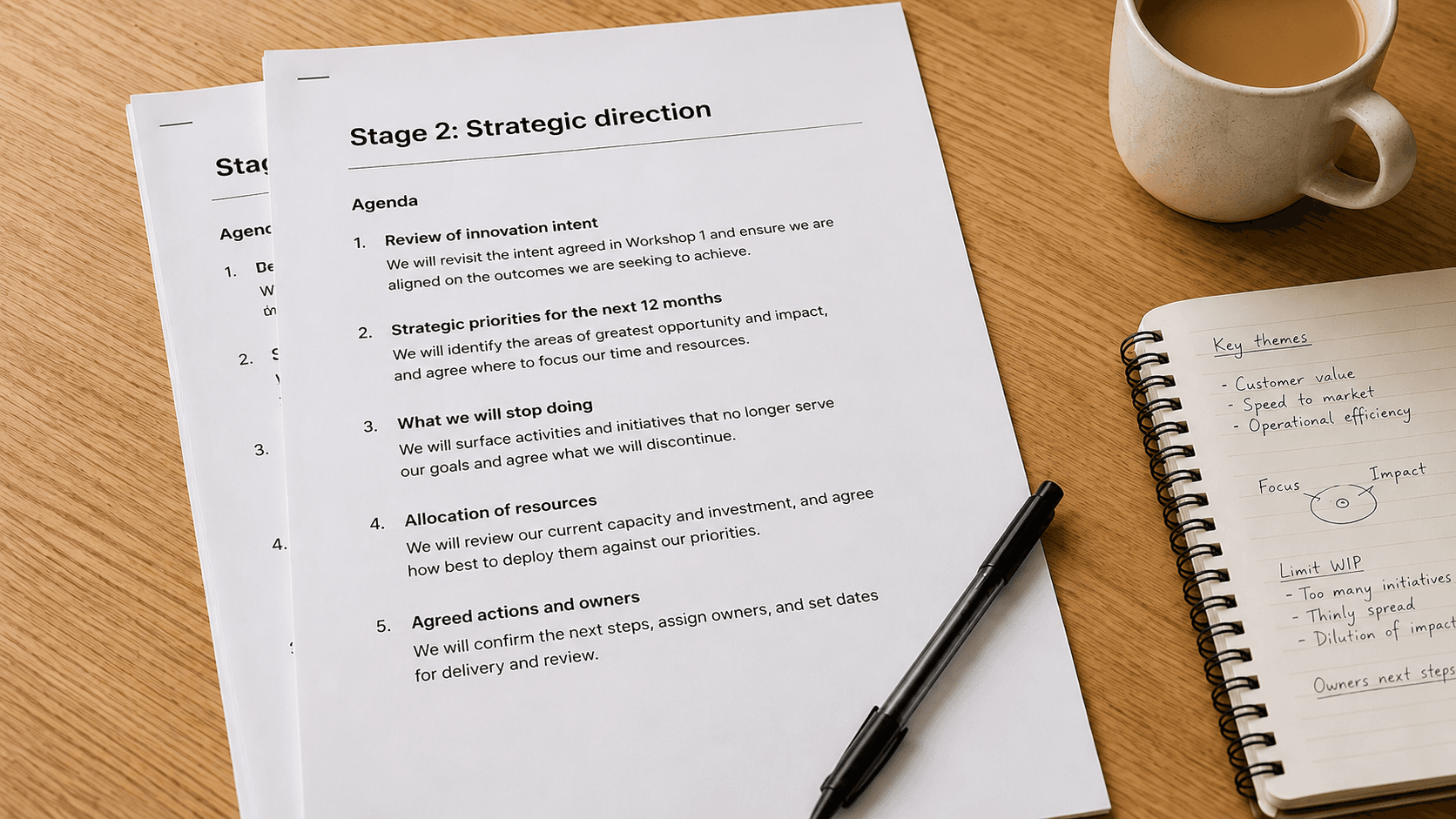 A working handout from a consulting engagement lying on a light oak desk. The handout is headed 'Stage 2: Strategic direction' and shows a numbered agenda with sub-items. A biro rests across the handout and a spiral-bound notebook sits to the right with handwritten notes and a hand-drawn matrix. A ceramic mug of coffee with milk sits in the upper right.